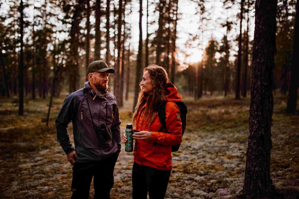 kaksi ihmistä metsässä nauttimassa toistensa seurasta, toisella termospullo kädessä, katsovat toisiaan hymyillen